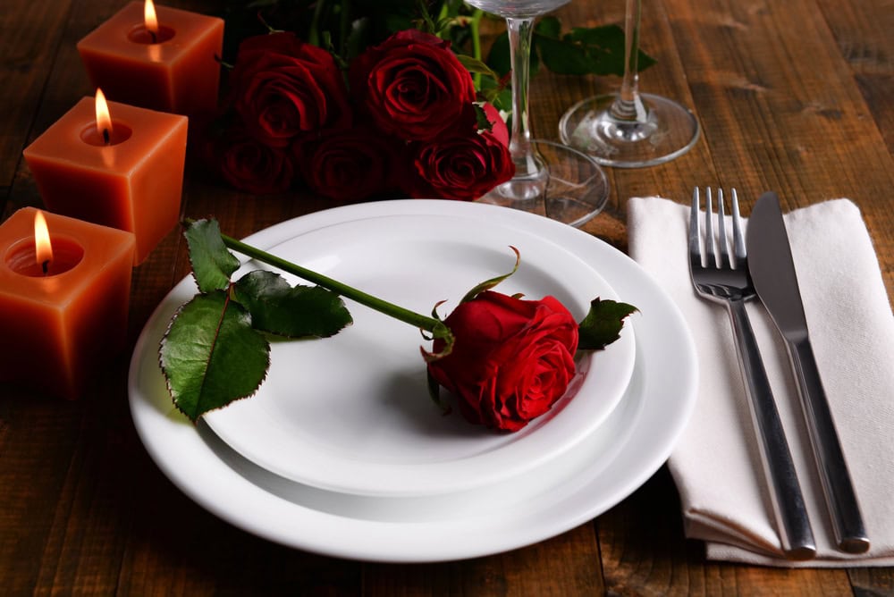 A table setting with a red rose on stacked white plates, silverware on a napkin, three lit candles, and a bouquet of roses on a wooden surface.