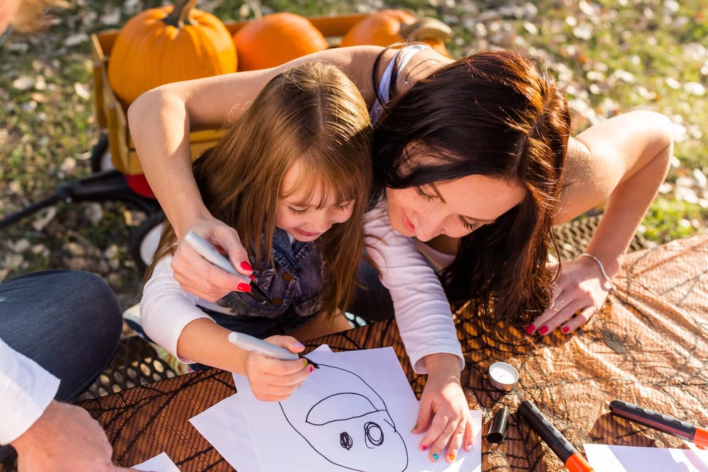 An adult helps a child draw on white paper at an outdoor table with pumpkins in the background. Markers and papers are scattered on the table.