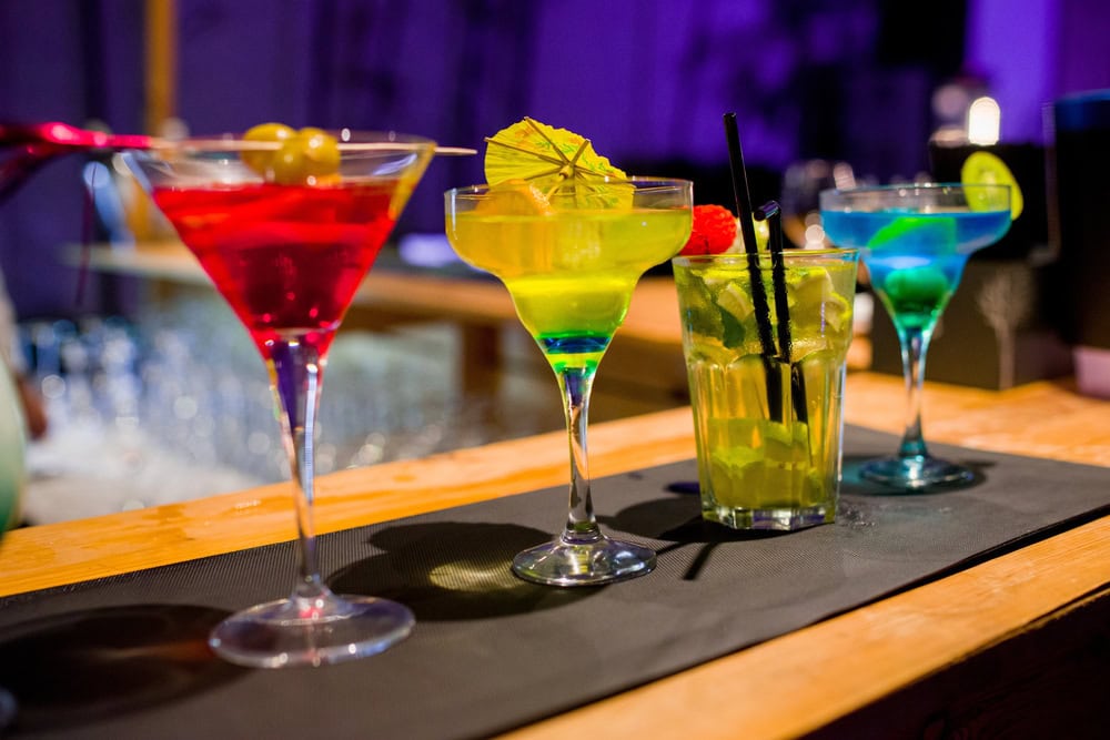 Four colorful cocktails in different glasses are lined up on a bar counter, each garnished with fruit or decorative items, against a blurred background.