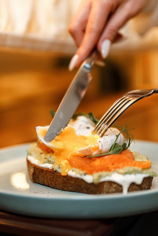A hand uses a knife and fork to cut into a poached egg on toast with smoked salmon and sauce, as the egg yolk runs onto the plate.