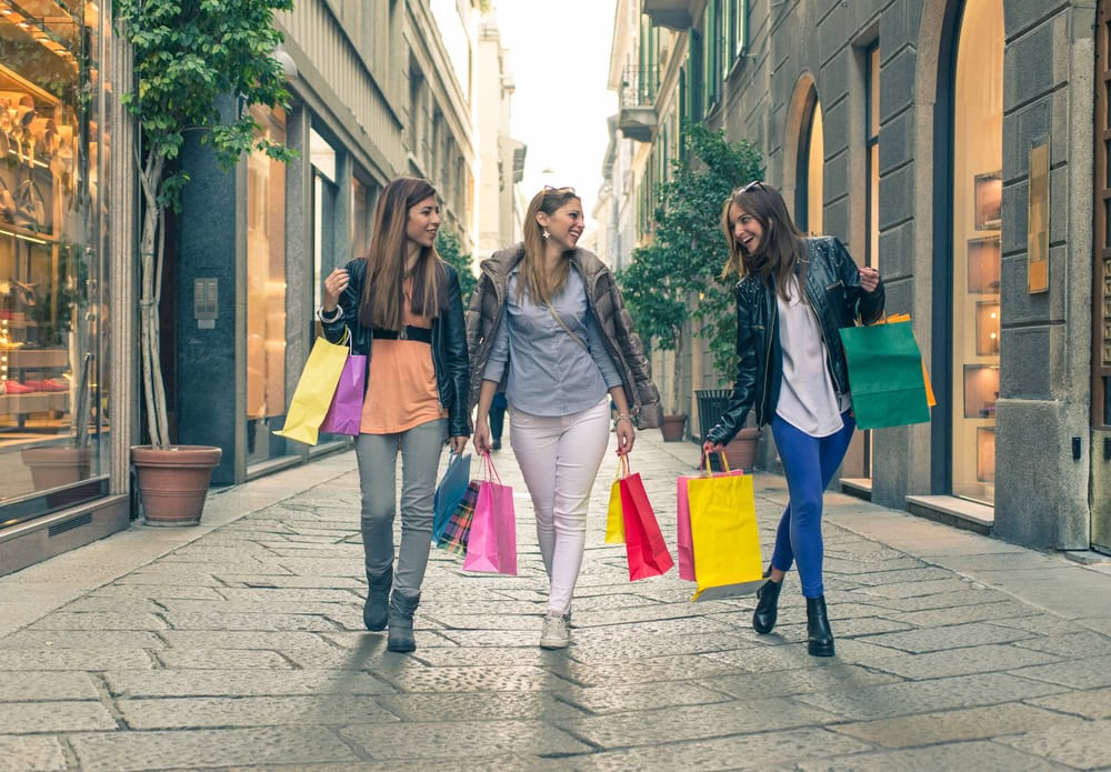 Three women walk down a shopping street carrying colorful shopping bags and smiling at each other.