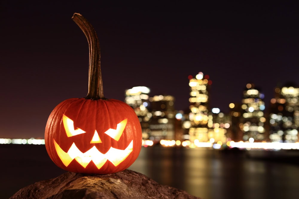 A carved jack-o’-lantern with a glowing face sits on a rock at night, with a city skyline and blurred lights in the background.