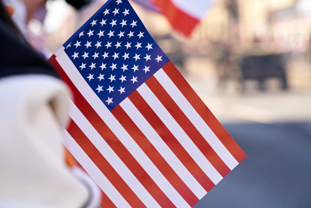 A close-up of a hand holding a small United States flag, with blurred background elements.