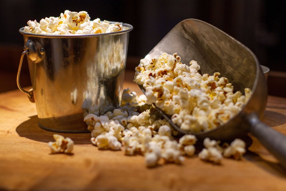 A metal bucket and a metal scoop filled with popcorn, with some popcorn spilled onto a wooden surface.