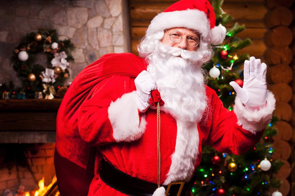 A person dressed as Santa Claus stands indoors by a Christmas tree and fireplace, holding a red sack and waving.