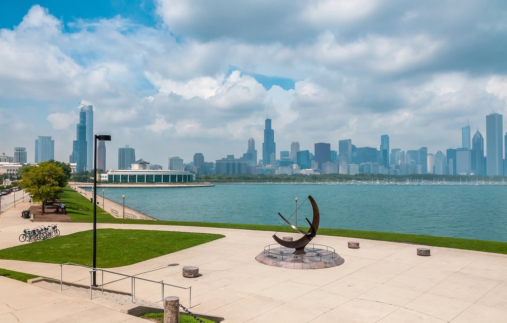 A lakeside walkway with a modern sundial sculpture in the foreground and the Chicago skyline in the background under partly cloudy skies.