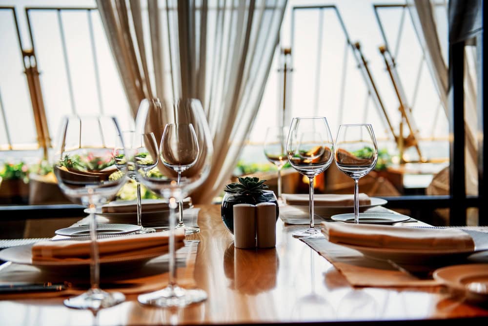 A neatly set dining table with wine glasses, plates, napkins, and a small potted plant in a sunlit restaurant with large windows and curtains.