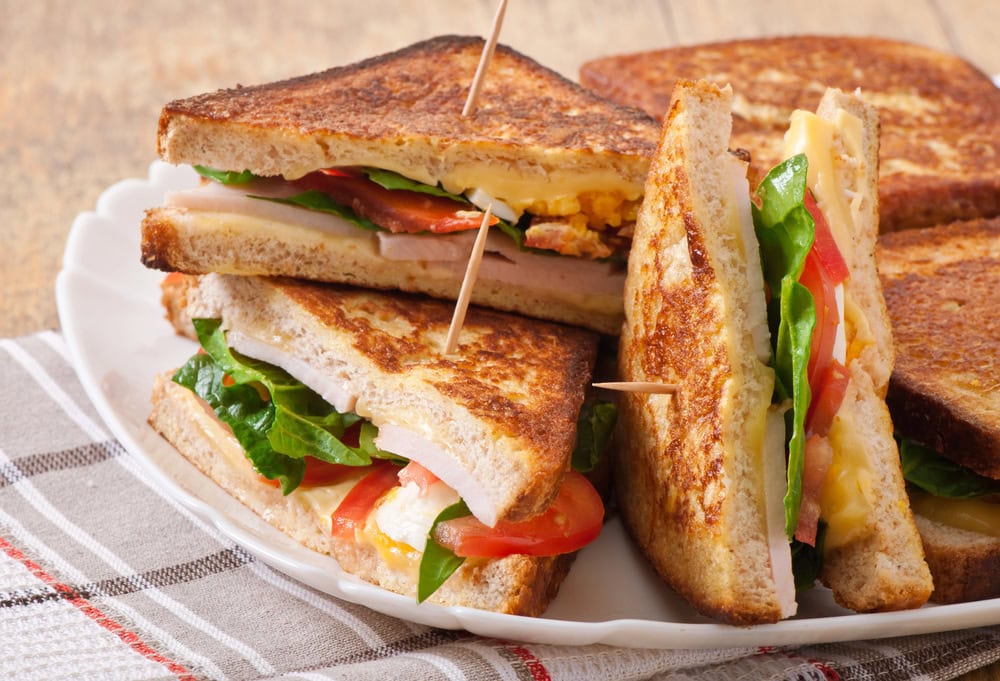 Close-up of club sandwiches cut in halves, stacked on a white plate with toothpicks, showing layers of bread, lettuce, tomato, cheese, ham, and egg.