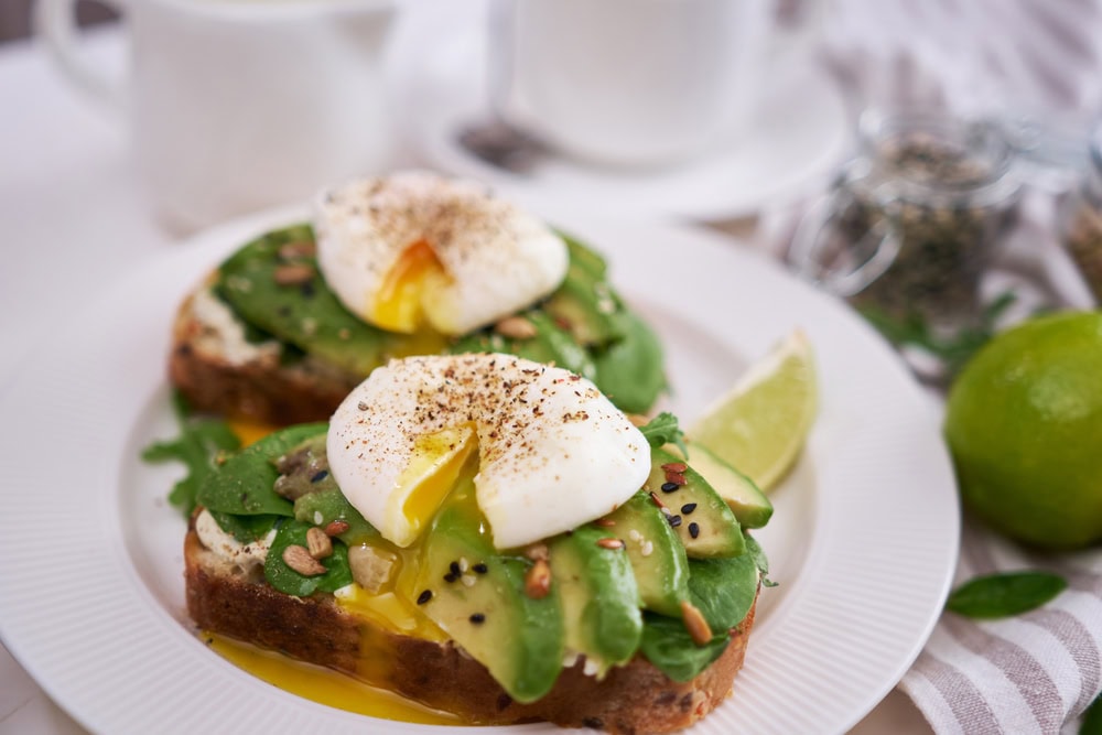 Two slices of toast topped with avocado, spinach, seeds, and poached eggs with runny yolks on a white plate, with a lime wedge and a blurred background.