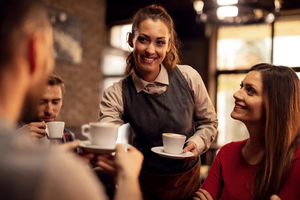 A waitress wearing an apron serves coffee to smiling customers at a café, with natural light coming through the windows in the background.
