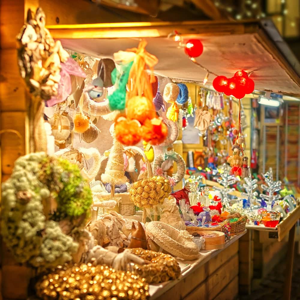 A market stall displays handmade crafts, including wreaths, ornaments, and decorations, all brightly lit and arranged on shelves and hanging from hooks.