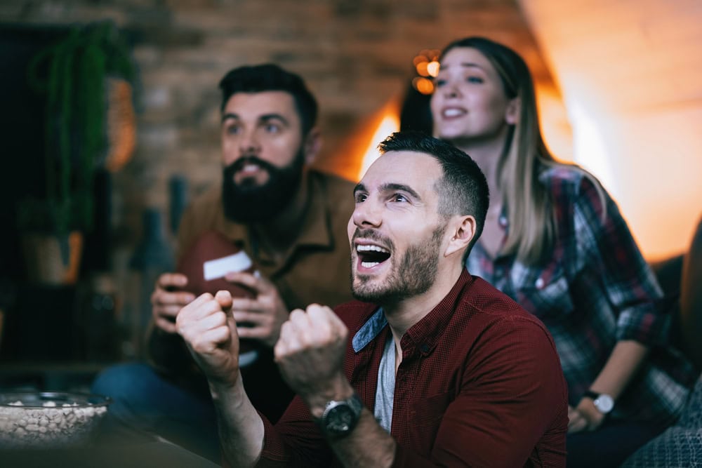 Three people sit indoors, watching something with excitement. The man in front is smiling and clenching his fists, while the two behind him also appear engaged.