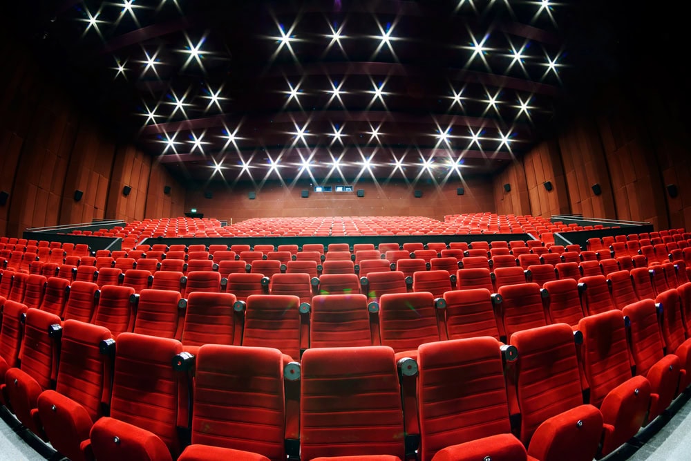 Wide-angle view of an empty theater auditorium with rows of red seats, a stage at the front, and ceiling lights illuminated above.