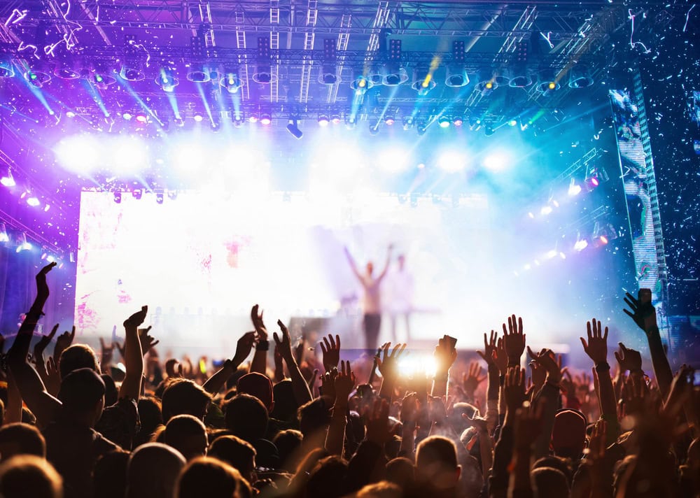 A large crowd with raised hands faces a brightly lit stage during a concert, with colorful lights and confetti in the air.