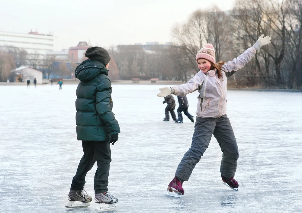 Two people ice skating outdoors on a frozen lake, with additional skaters and winter trees in the background. One person is posing with arms outstretched.