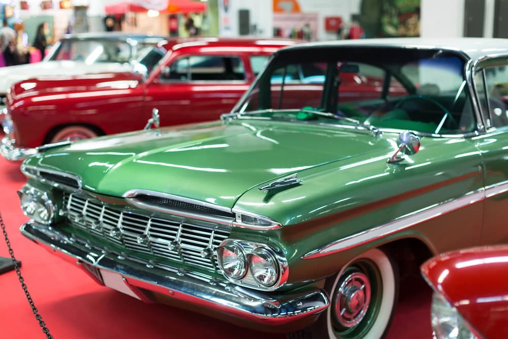 A green classic car is displayed indoors alongside other vintage vehicles on a red carpet at an automotive show.
