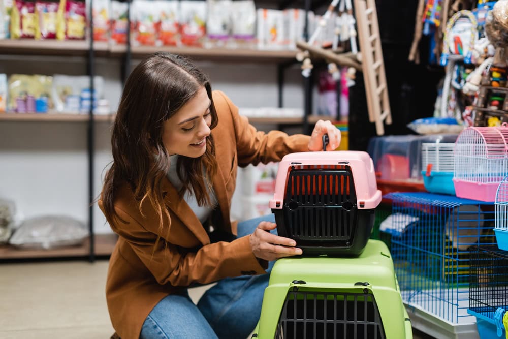 A woman in a pet store kneels and examines a pink and black pet carrier stacked on a green one, surrounded by cages and pet supplies.