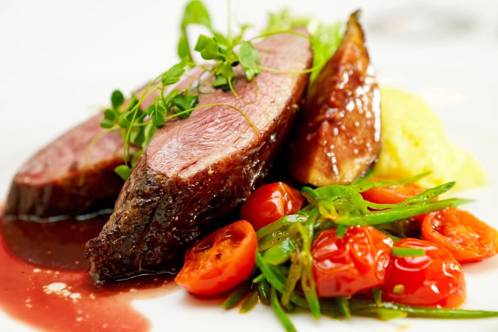 Sliced medium-rare steak with brown sauce, mashed potatoes, cherry tomatoes, snap peas, and greens on a white plate.