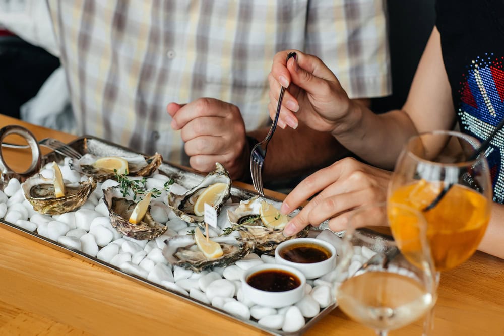 Two people seated at a table eat fresh oysters with lemon wedges and sauce, accompanied by drinks.