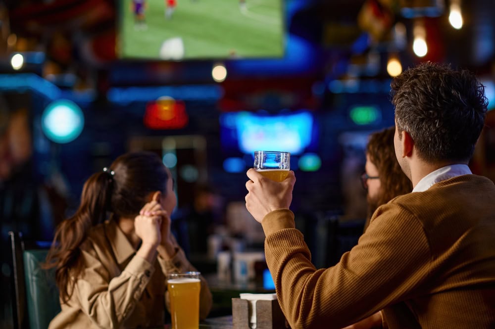 Three people sit at a bar table, two holding glasses of beer, watching a sports game on a large TV screen in the background.