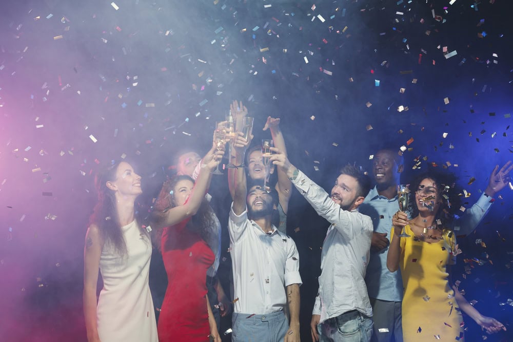 A group of people in colorful outfits raise glasses in a toast as confetti falls around them, celebrating under dramatic lighting.
