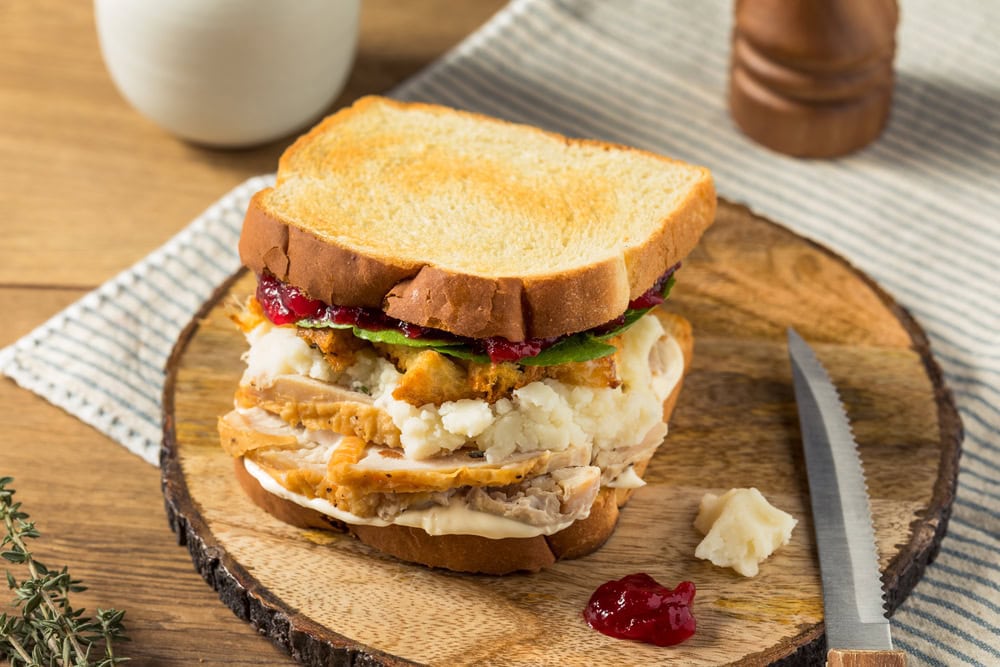 A sandwich with turkey, stuffing, mashed potatoes, gravy, lettuce, and cranberry sauce on toasted bread sits on a wooden plate with a knife beside it.