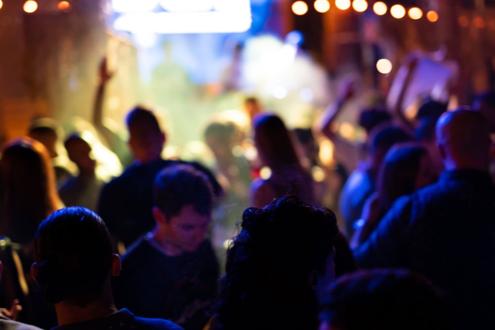 A crowd of people stands and dances in a dimly lit venue with colorful lights and smoke, creating a lively party atmosphere.