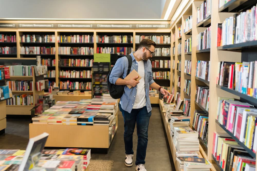 A man wearing glasses and a backpack browses books on a shelf in a well-lit bookstore, holding a book in one hand.