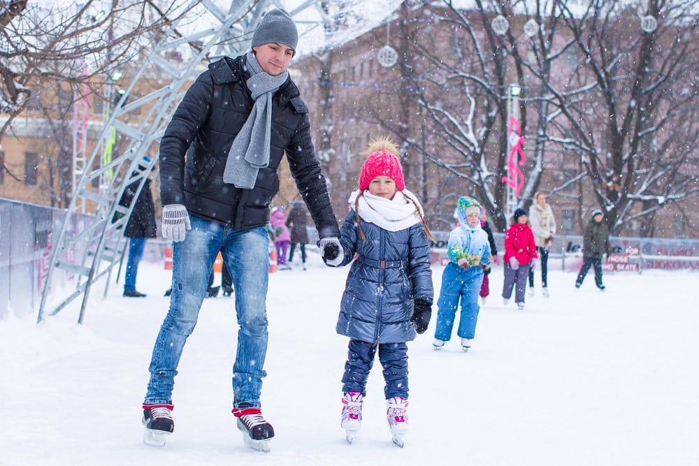 A man and a young girl ice skating outdoors on a snowy day, holding hands, with other people skating in the background.