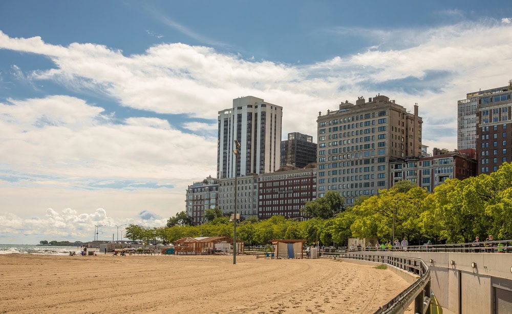 A sandy beach with a few people, bordered by a walkway, green trees, and tall buildings under a partly cloudy sky.