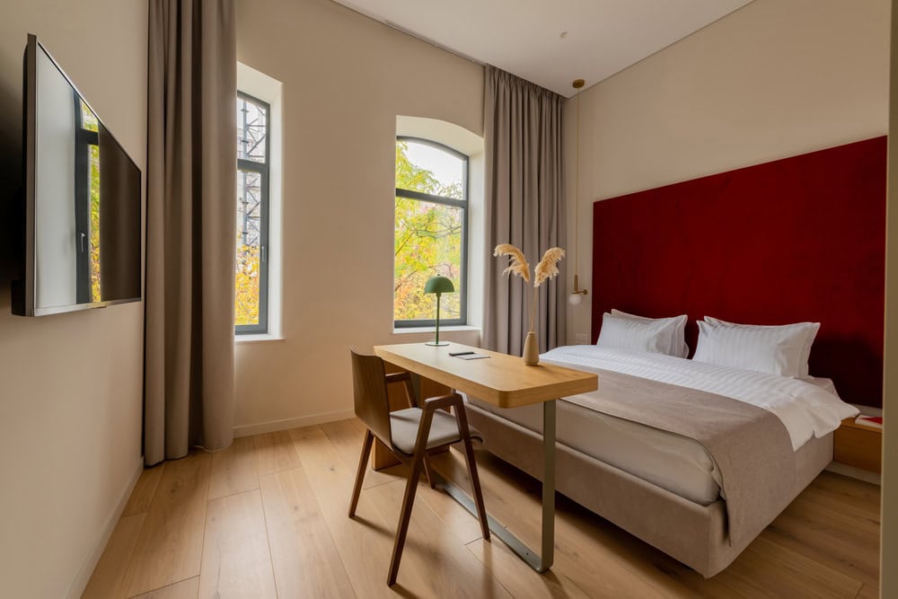 Modern hotel room with a double bed, red headboard, wall-mounted TV, wooden desk and chair, large windows with curtains, and neutral decor.