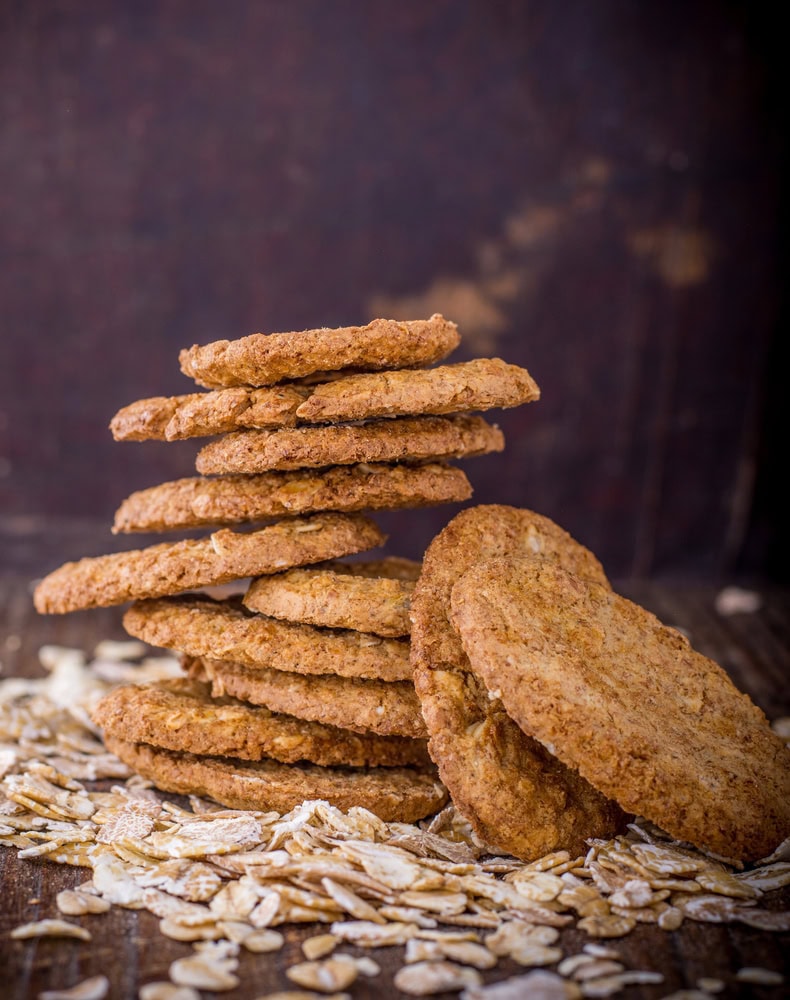 A stack of oat cookies sits on a wooden surface surrounded by scattered oats, with two cookies leaning against the stack.