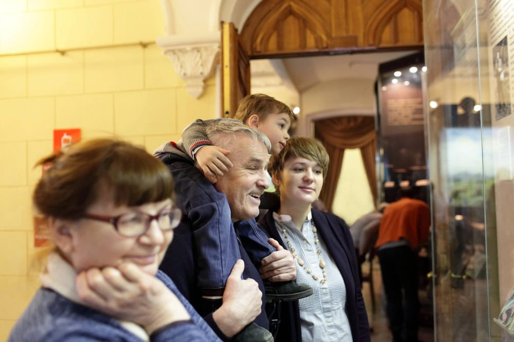 Four people, including a child on an older man's shoulders, look at a museum display case. Two women stand nearby, and the group appears engaged in observing the exhibit.