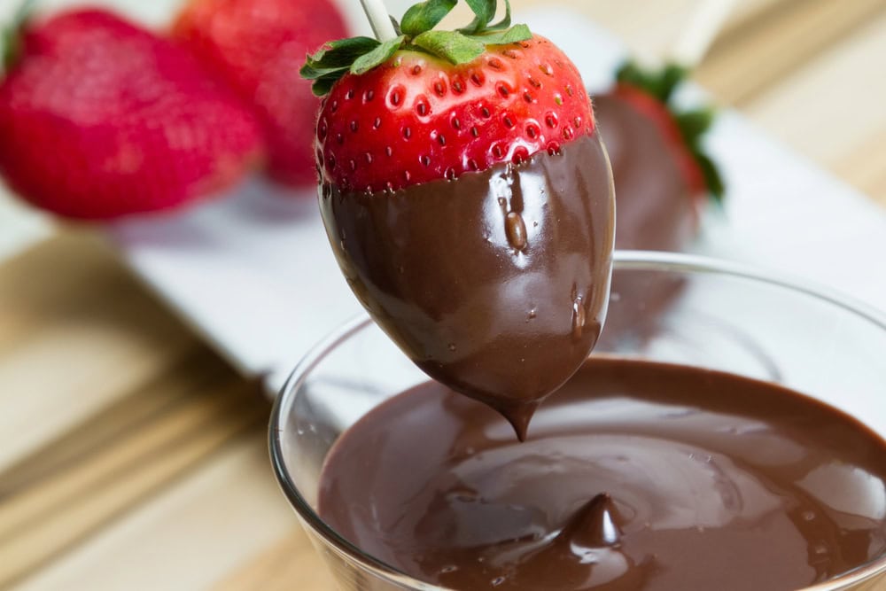 A fresh strawberry being dipped into a bowl of melted chocolate, with more strawberries and chocolate in the background.