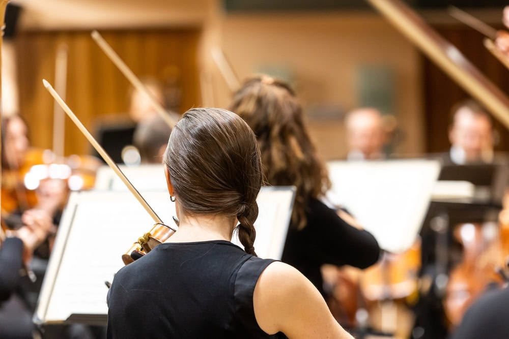 Musicians with string instruments rehearse in an orchestra, focusing on sheet music in a well-lit concert hall.