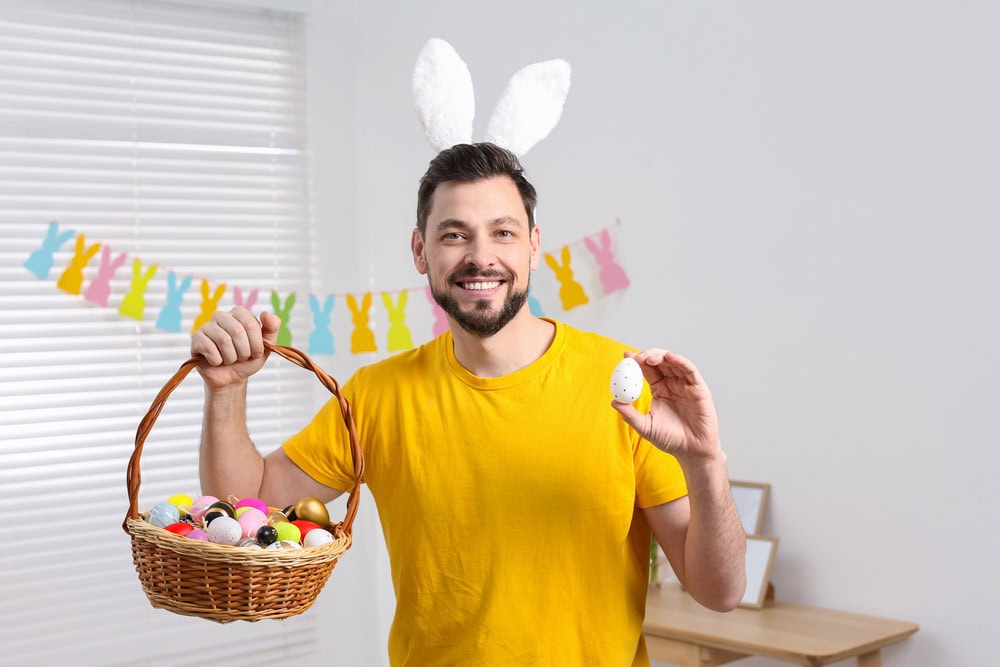 Man wearing bunny ears and a yellow shirt holds a basket of colorful Easter eggs in one hand and a single egg in the other, standing indoors with bunny decorations in the background.