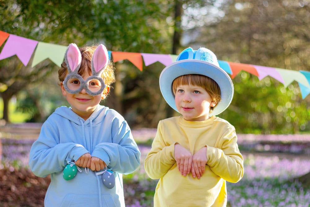 Two children in pastel outfits pose outdoors, wearing bunny ears and a blue hat, with colorful bunting and springtime flowers in the background.