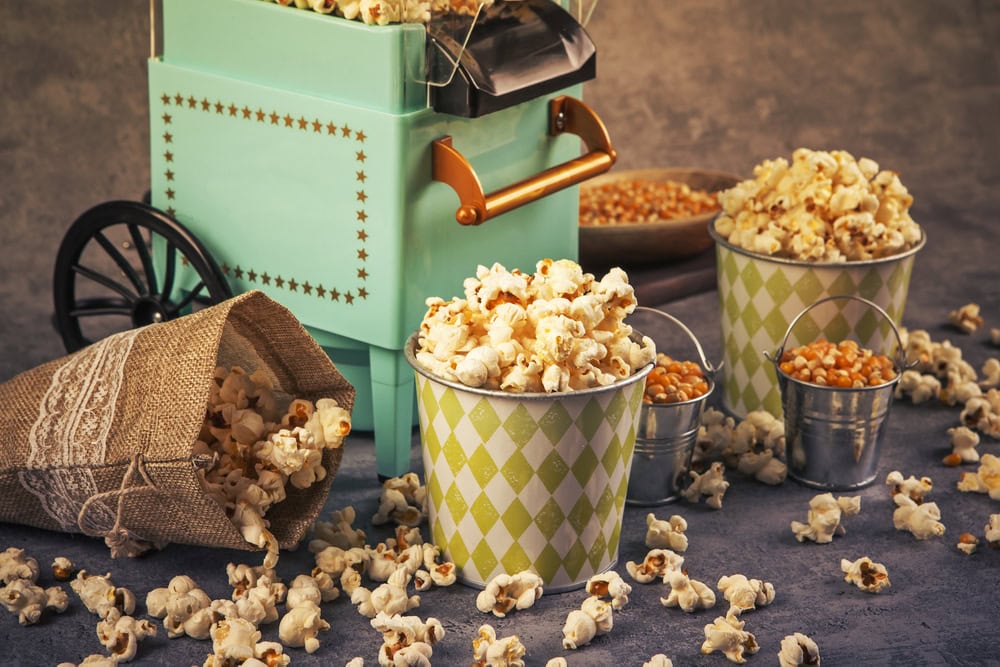 Vintage-style popcorn machine with popcorn overflowing from cups and a burlap sack, surrounded by scattered popcorn and small buckets of corn kernels on a textured surface.