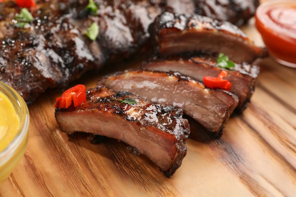 Sliced barbecued pork ribs with glaze, garnished with small red chili pieces, served on a wooden board with sauces nearby.