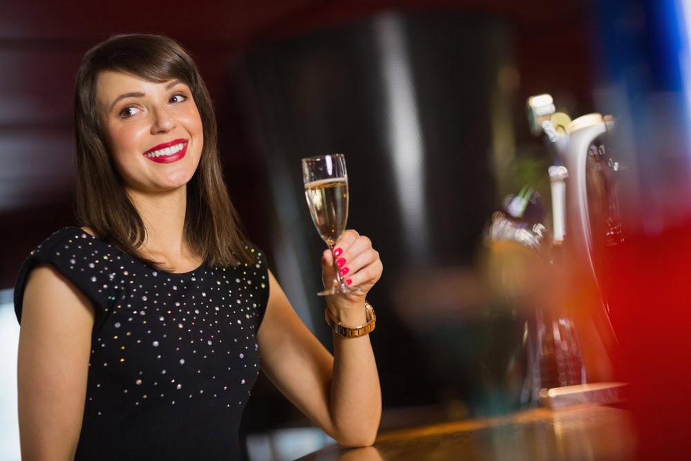 Woman with brown hair wearing a black, beaded top holds a glass of champagne and smiles while sitting at a bar.