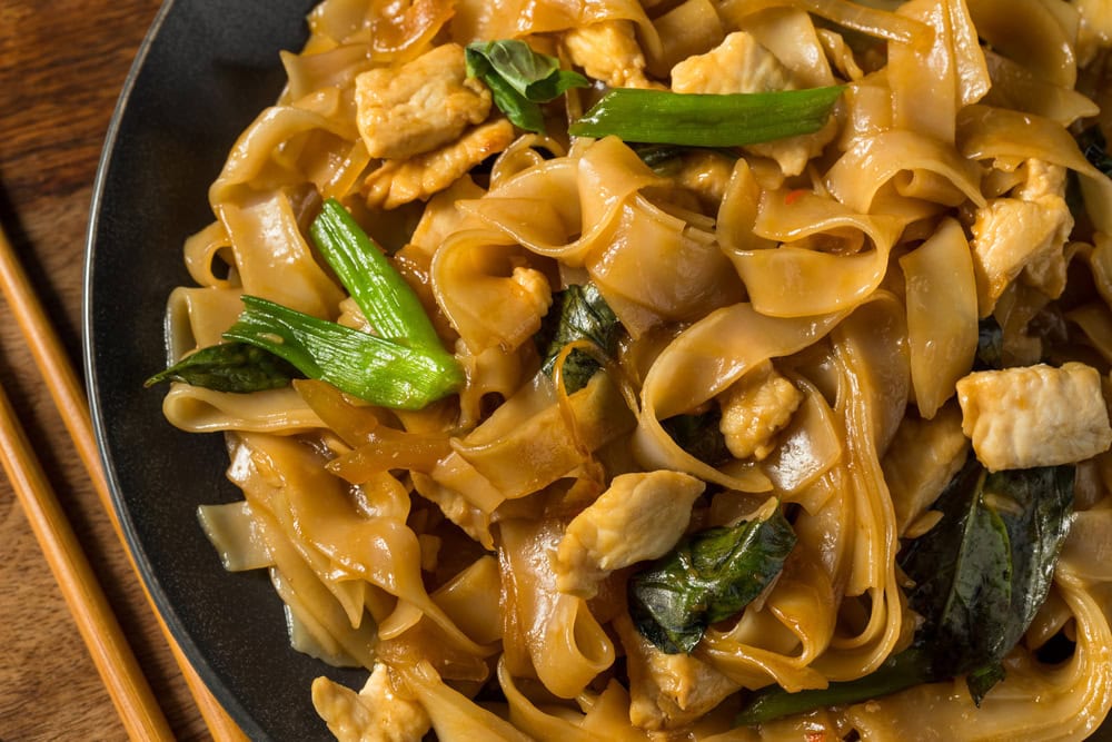 A plate of stir-fried flat rice noodles with pieces of chicken, green vegetables, and basil, served with chopsticks.