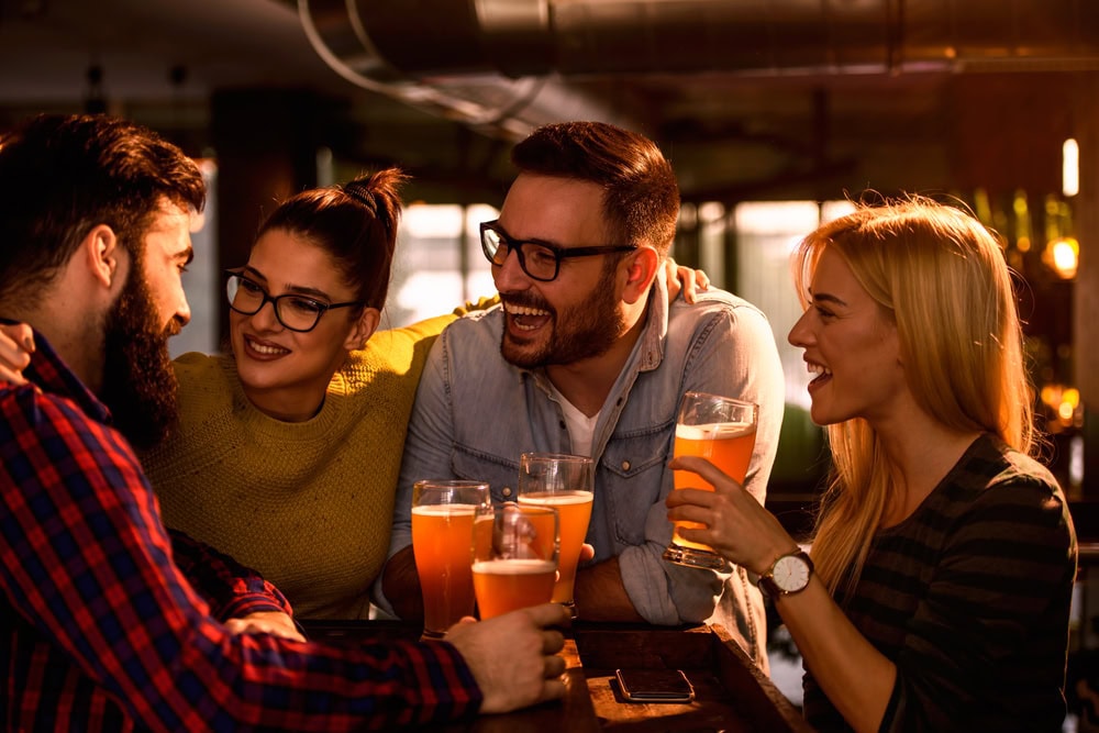 Four people sit at a bar table, smiling and talking while holding glasses of beer.