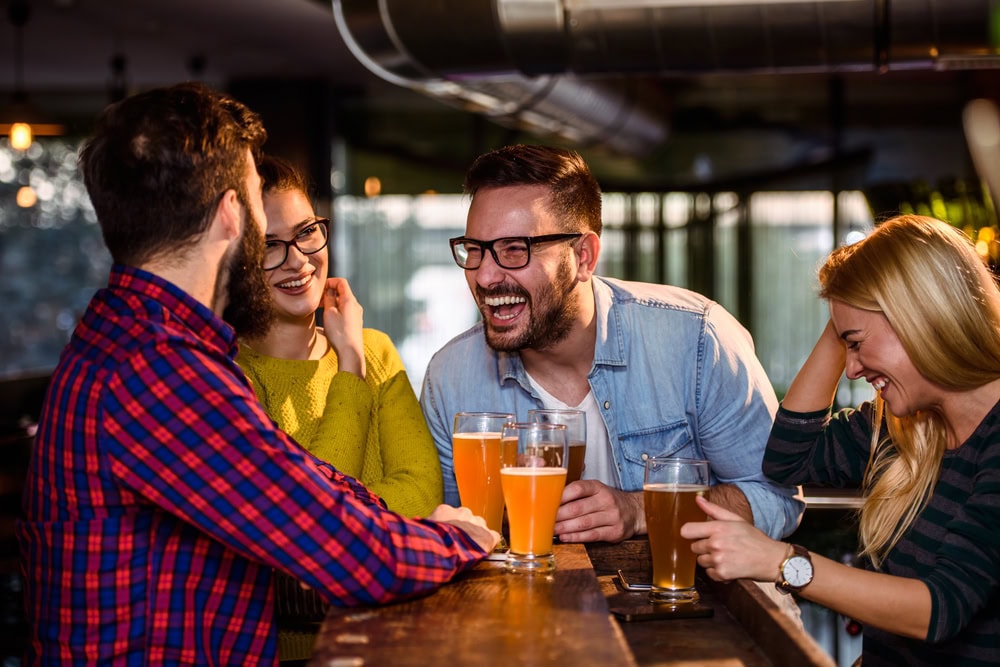 Four people sit at a bar table with beer and cocktails, laughing and talking together in a casual, indoor setting.
