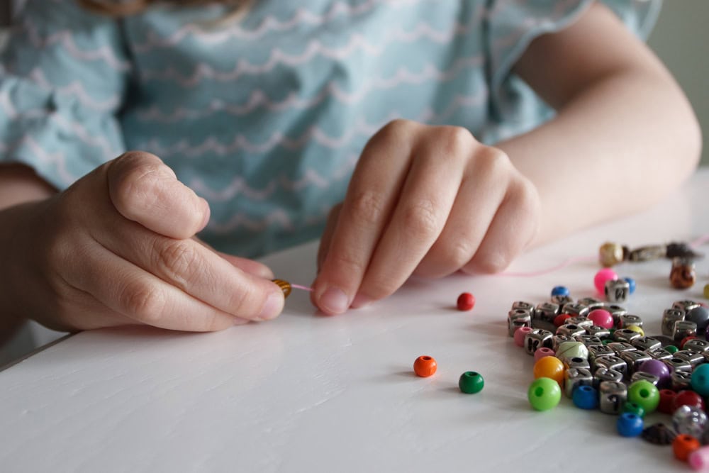 A child’s hands thread a bead onto string at a table, with assorted colorful beads scattered nearby.