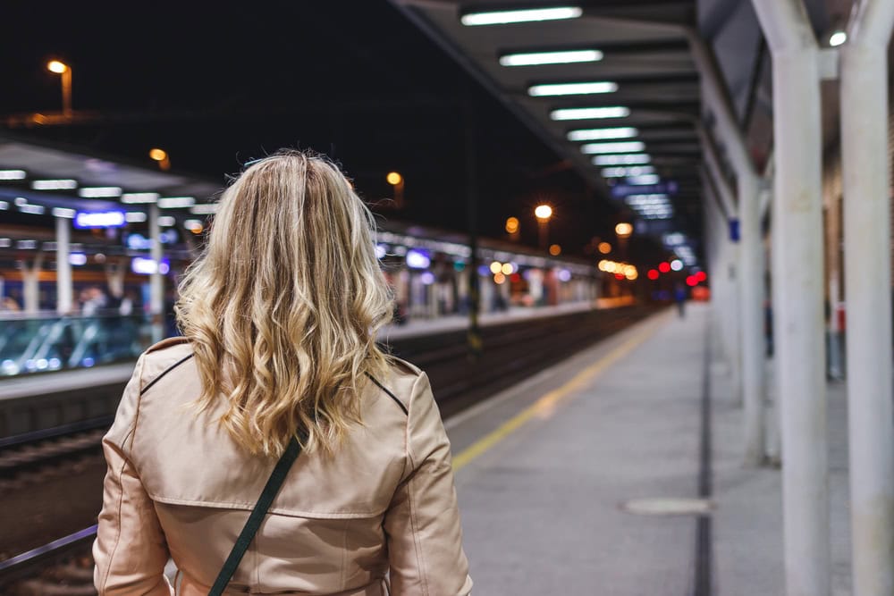 A woman with blonde hair and a beige coat stands on a train platform at night, facing the tracks and station lights.