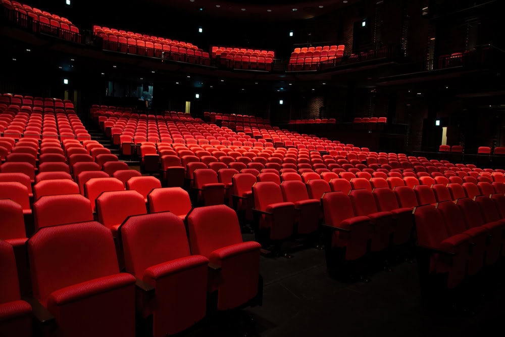 Rows of empty red theater seats in a dark auditorium, with lights on along the walls and aisles.