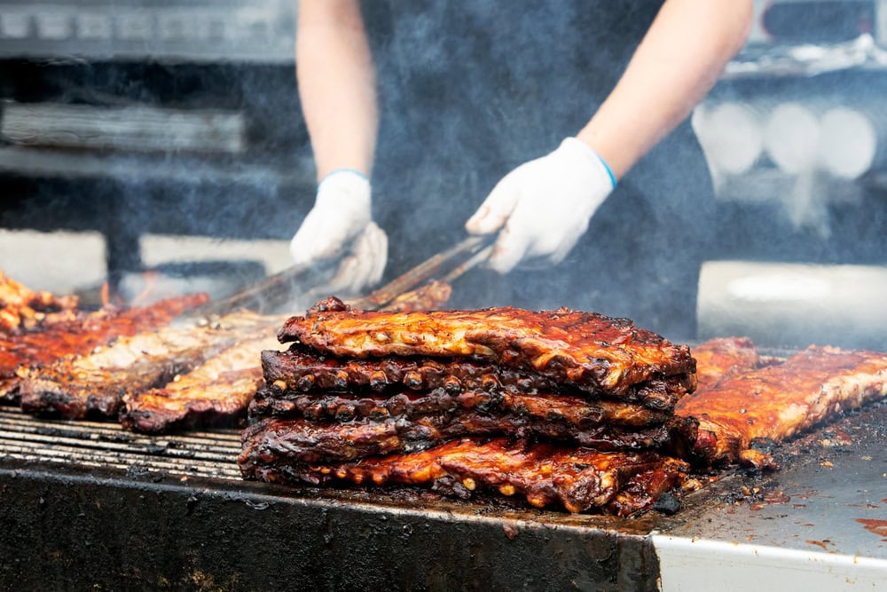 A person wearing gloves cooks and stacks racks of barbecued ribs on a grill with smoke rising in the background.