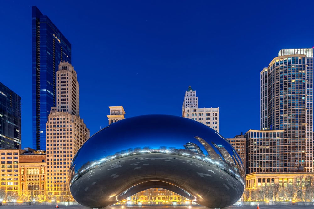 Cloud Gate sculpture, also known as "The Bean," in Chicago's Millennium Park at night, with surrounding city buildings illuminated.