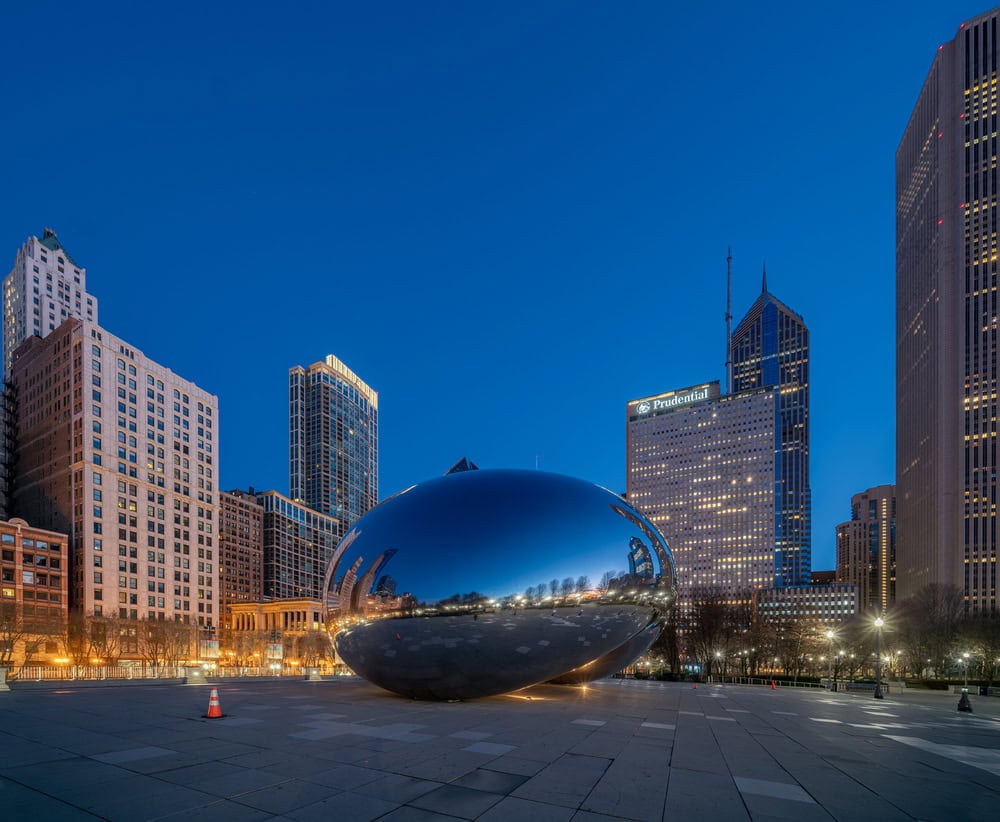 A large reflective sculpture, known as "The Bean," sits in a city plaza surrounded by tall buildings at dusk.