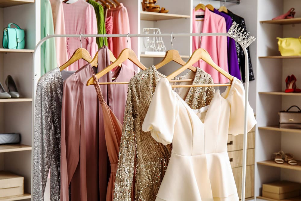 Four elegant dresses, including a white puff-sleeve gown and a gold sequin dress, hang on a rack in a walk-in closet with shelves holding shoes, bags, and folded clothes.